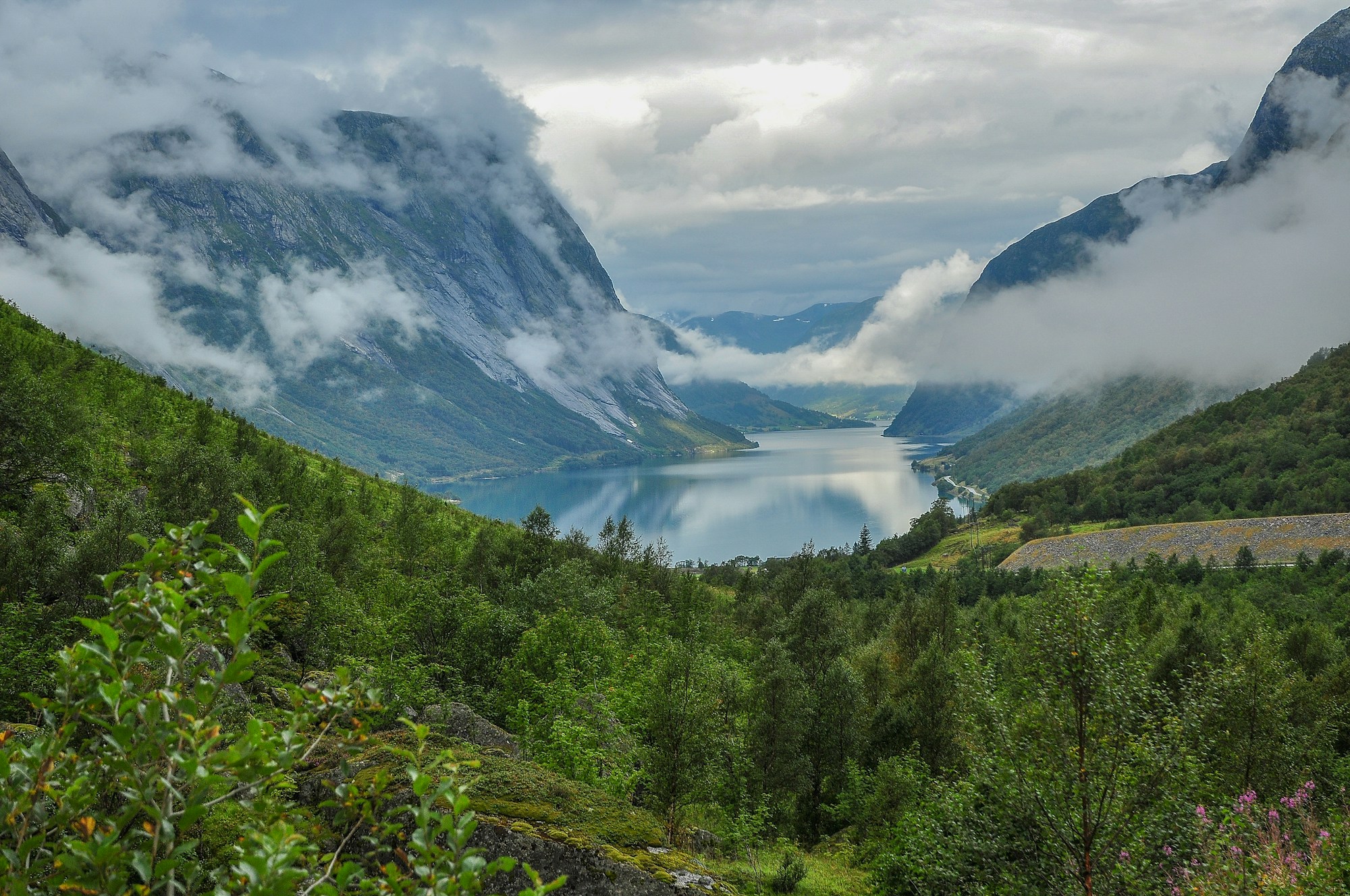 A quiet Norwegian fjord between green mountain slopes and low drifting cloud